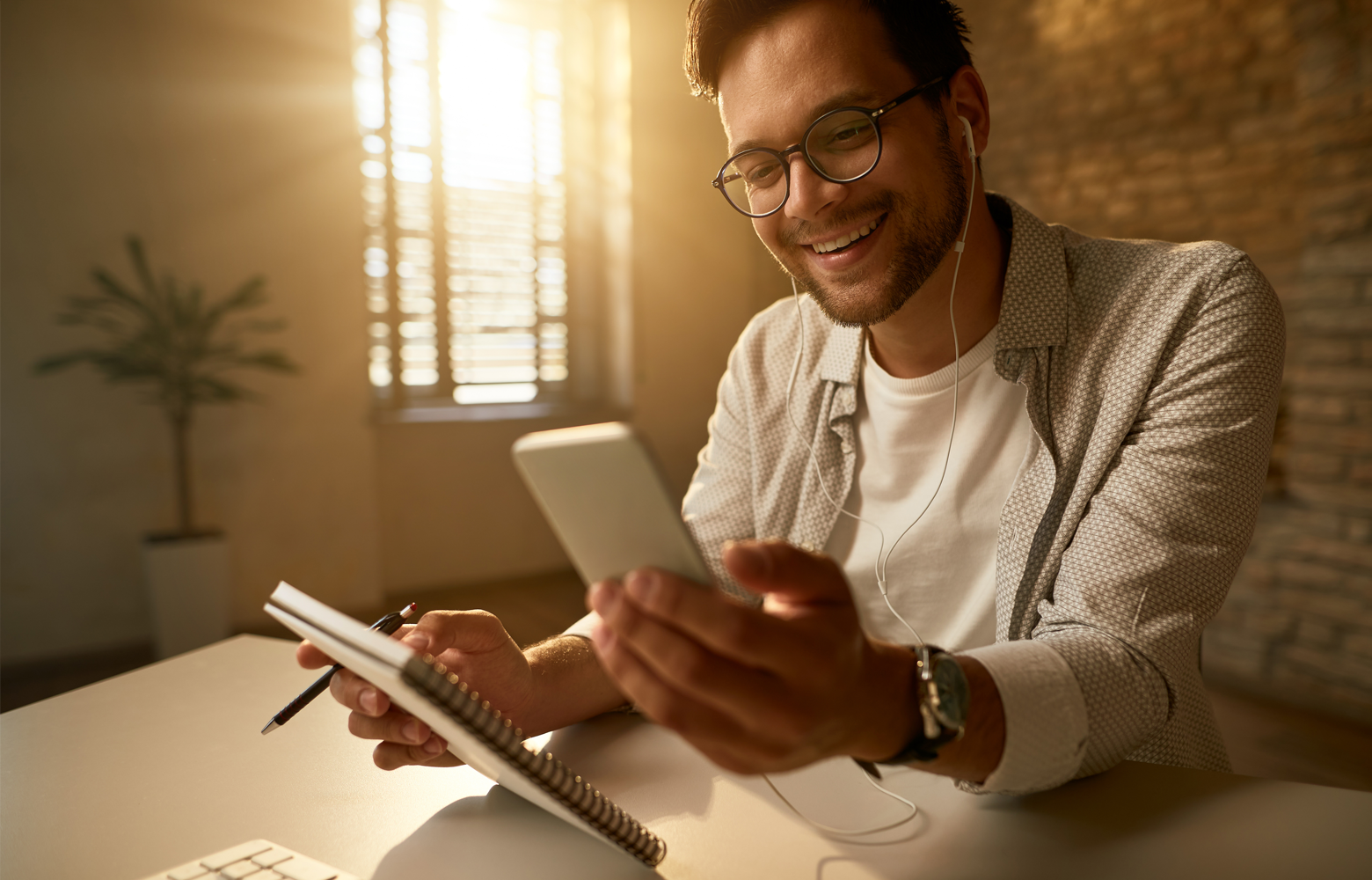 Homem sorrindo enquanto utiliza o celular e faz anotações em um caderno, em ambiente de trabalho, representando a criação de conteúdo.