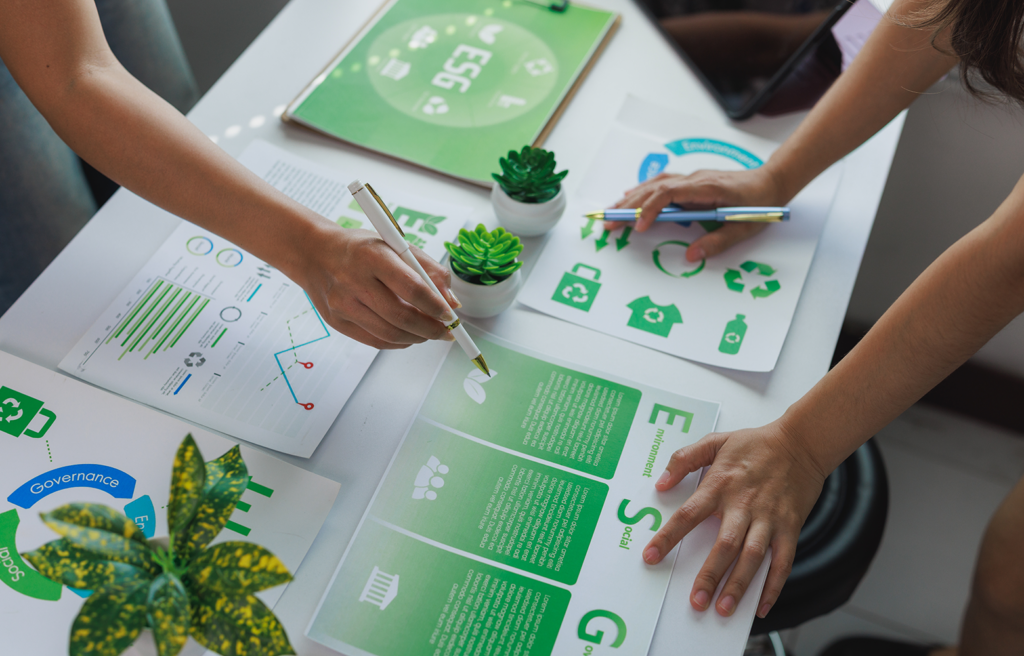 Equipe analisando relatórios e indicadores ESG em uma mesa de trabalho, com gráficos, ícones de reciclagem e materiais relacionados à sustentabilidade. A imagem representa práticas sustentáveis aplicadas à gestão empresarial e à geração de valor para as marcas.