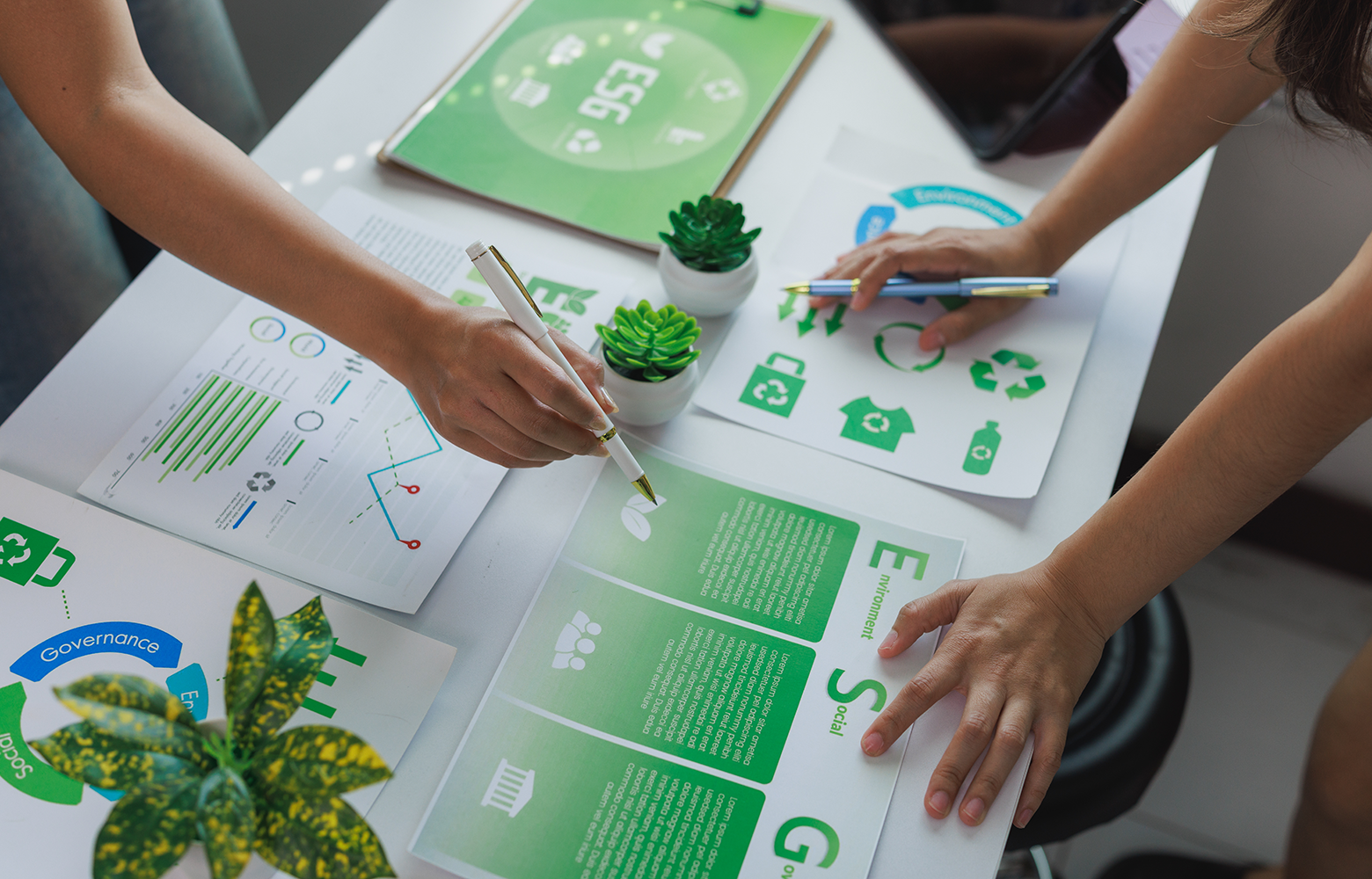 Equipe analisando relatórios e indicadores ESG em uma mesa de trabalho, com gráficos, ícones de reciclagem e materiais relacionados à sustentabilidade. A imagem representa práticas sustentáveis aplicadas à gestão empresarial e à geração de valor para as marcas.