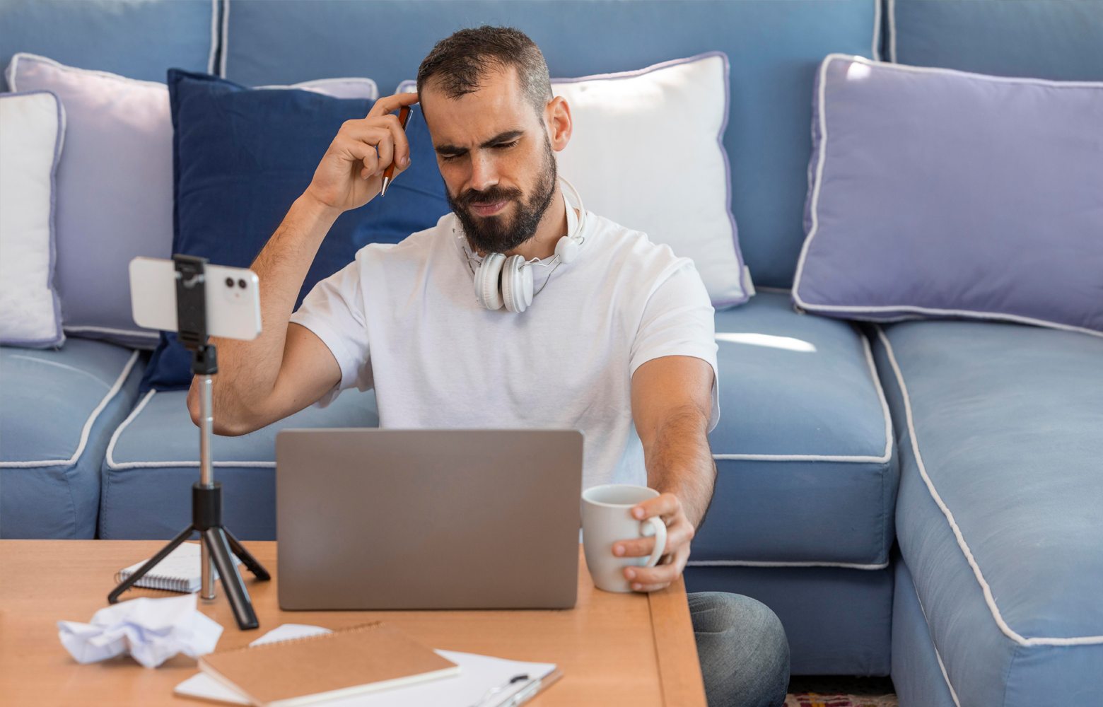 Empreendedor sentado em frente a um notebook e a um celular apoiado em tripé, refletindo sobre a criação de conteúdo para a internet em um ambiente de trabalho doméstico.
