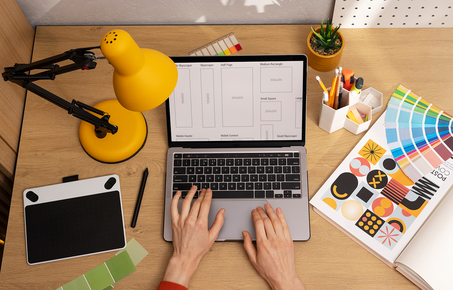 Profissional trabalhando em notebook em uma mesa de design com amostras de cores, tablet gráfico e materiais criativos, representando a construção de uma identidade visual estratégica para marcas.