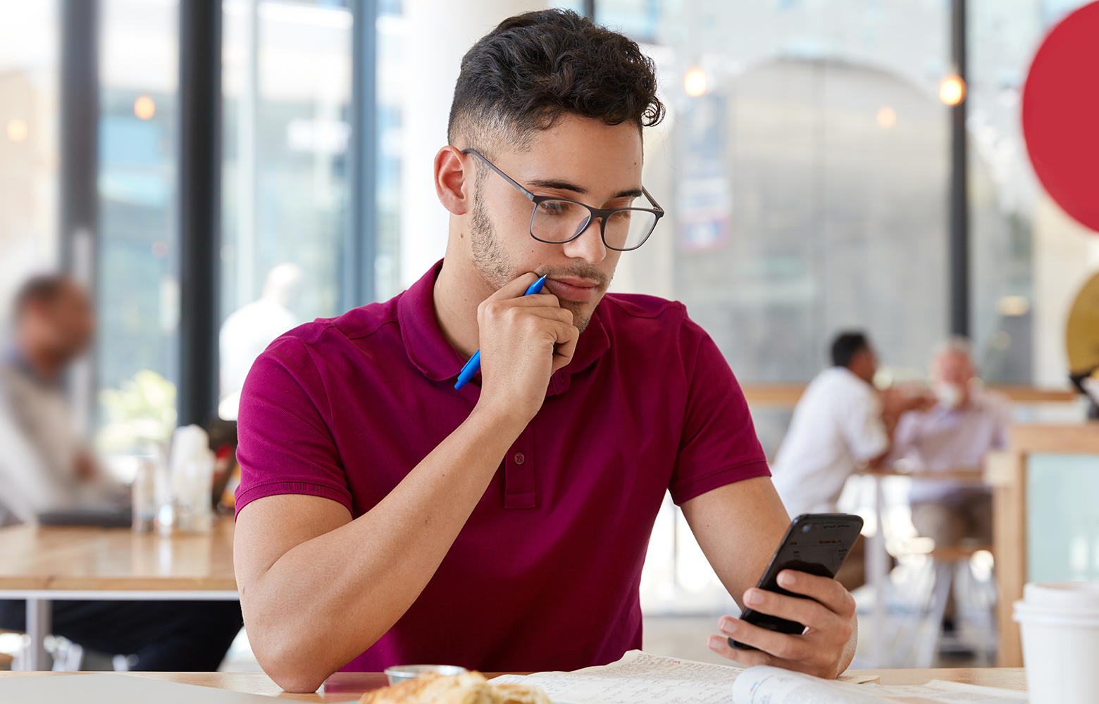Pessoa em uma cafeteria pensando enquanto observa informações no celular enquanto faz anotações. A imagem representa o processo de pesquisa e decisão do consumidor, destacando a importância da lembrança de marca para que empresas sejam recordadas no momento da escolha.