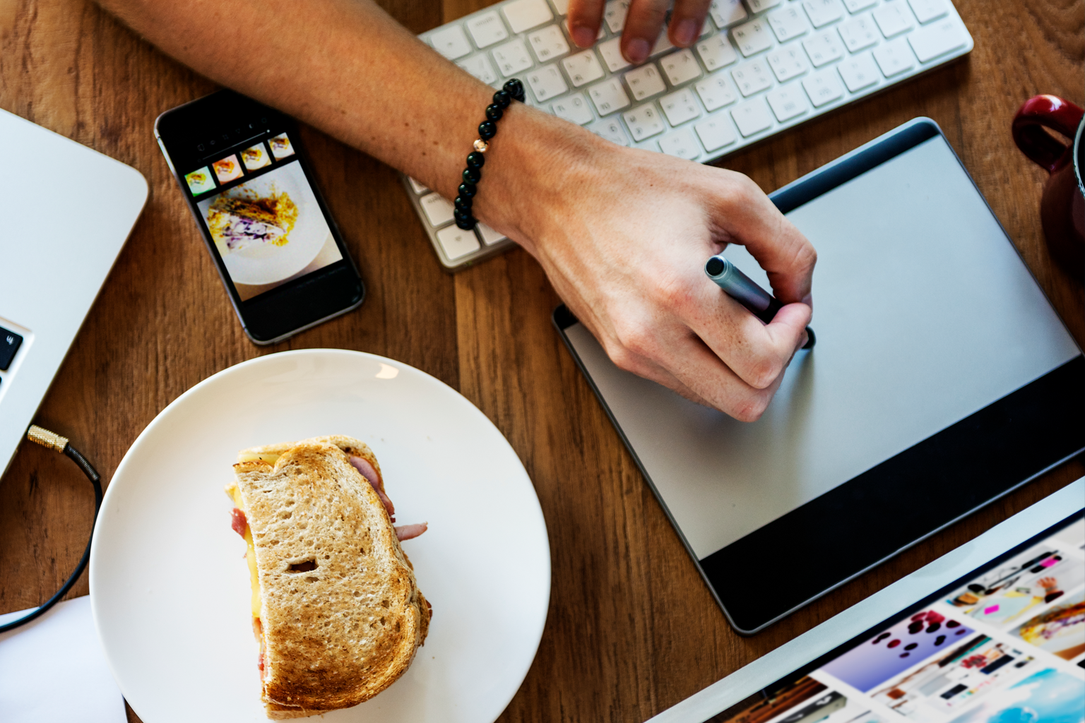 Profissional cria conteúdo digital em uma mesa de trabalho com tablet gráfico, celular e computador, enquanto faz uma pausa para comer. A imagem ilustra a produção de conteúdo para estratégias de SEO, encontrabilidade e conversão de visitantes em clientes.